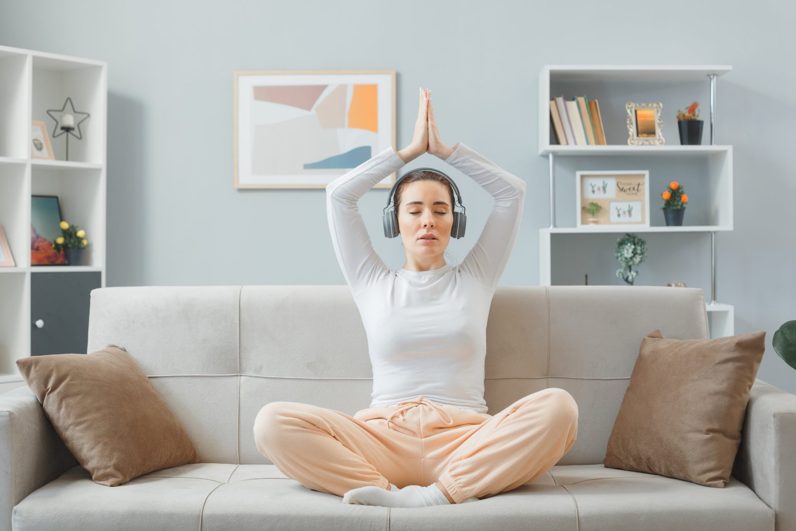 young beautiful woman in home clothes sitting on a couch at home interior with headphones happy and positive relaxing meditating making meditation gesture holding palms together over her head spending weekend at home
